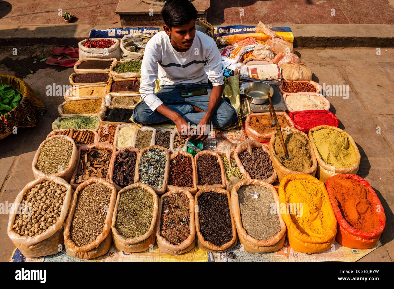 Un commerciante siede a gambe incrociate in un mercato di Varanasi e offre un'ampia varietà di spezie aromatiche e polveri ordinate in sacchi aperti. Varanasi UTT Foto Stock