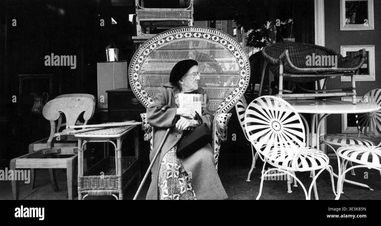 A partire dagli anni '1950, questa foto di strada di Angelo Rizzuto cattura una donna anziana seduta comodamente su una grande sedia in vimini pavone fuori da un negozio di mobili a New York. Ha un giornale, la sua borsa e un bastone, che incarna un momento di tregua e di vita quotidiana in una strada di città. Foto Stock