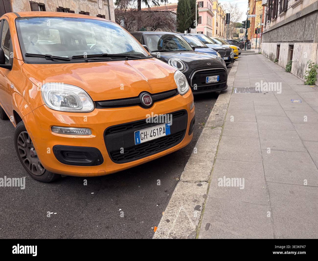 Primo piano di una Fiat Panda arancione parcheggiata accanto a uno stretto marciapiede in pietra a Roma, Italia, con altre auto compatte in coda. Trasporto urbano di tutti i giorni. Foto Stock