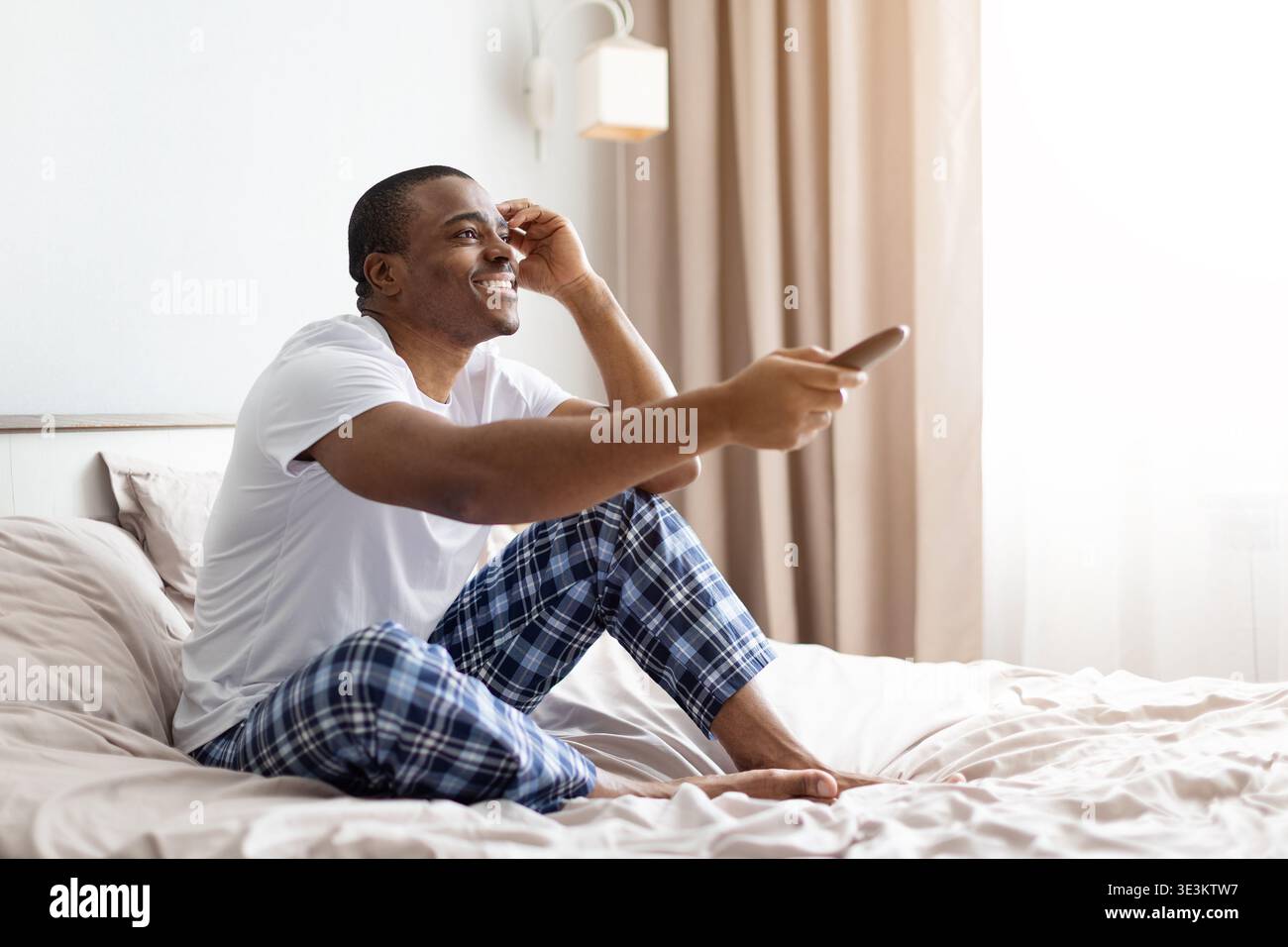 Giovane uomo nero seduto sul letto sorridente tenendo il telecomando Foto Stock