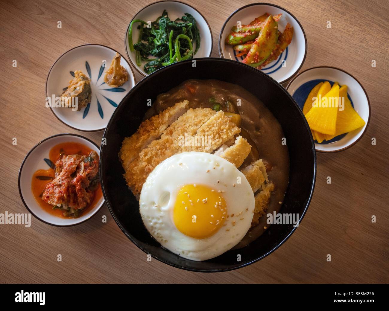 Katsu curry coreano di fusione giapponese con croccante cotoletta di maiale impanata, riso e uova fritte, servito con contorni coreani assortiti su un tavolo di legno Foto Stock