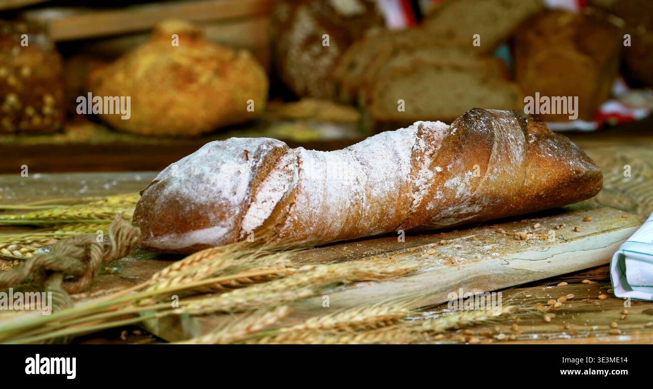 Pane appena sfornato su una tavola di legno con grano e altri tipi di pane in un ambiente da forno Foto Stock