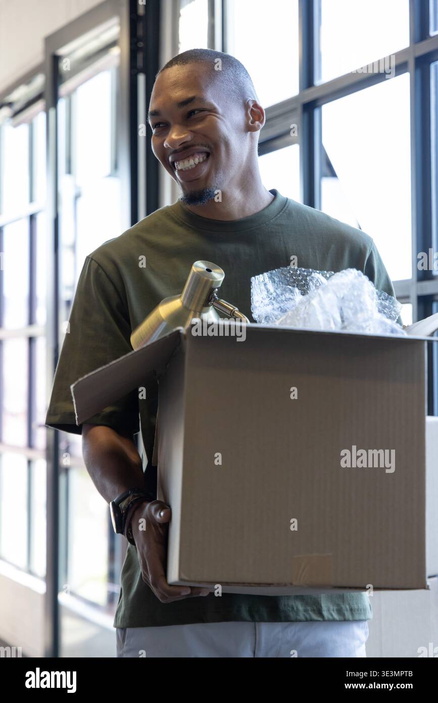 Uomo afro-americano che tiene in mano una scatola mobile con bolla avvolgente, disimballando la lampada in ottone vicino alle finestre della griglia Foto Stock