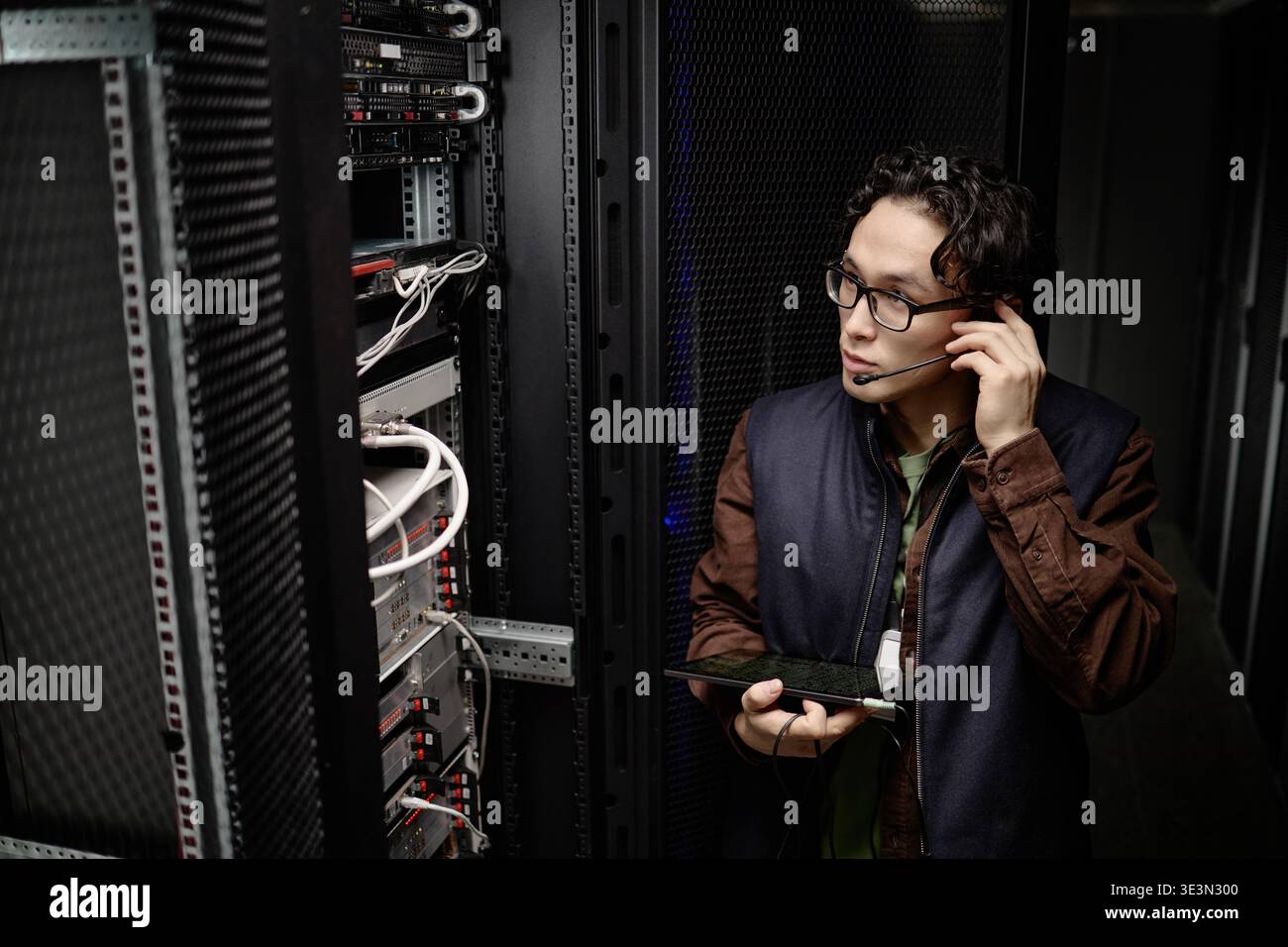 Giovane uomo asiatico che monitora il rack server durante l'utilizzo di un tablet digitale Foto Stock
