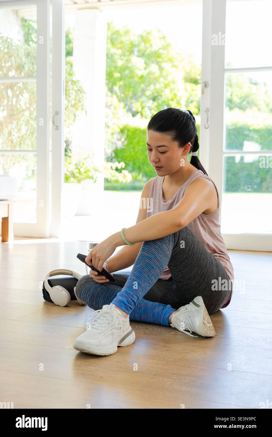 Donna asiatica seduta su un pavimento di legno vicino a porte con le cuffie per smartphone che indossano leggings tanktop Foto Stock