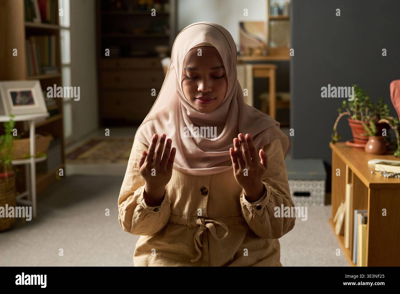 Donna musulmana adulta seduta in casa alzando le mani in preghiera, indossando hijab, occhi chiusi, praticando rituali religiosi, circondata da elementi interni e librerie Foto Stock