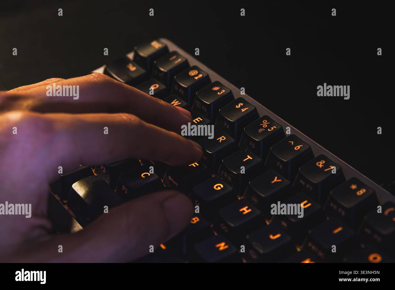 Un primo piano dettagliato della mano di una persona che preme i tasti su una moderna tastiera meccanica per computer. I tasti sono dotati di una calda retroilluminazione arancione. Foto Stock