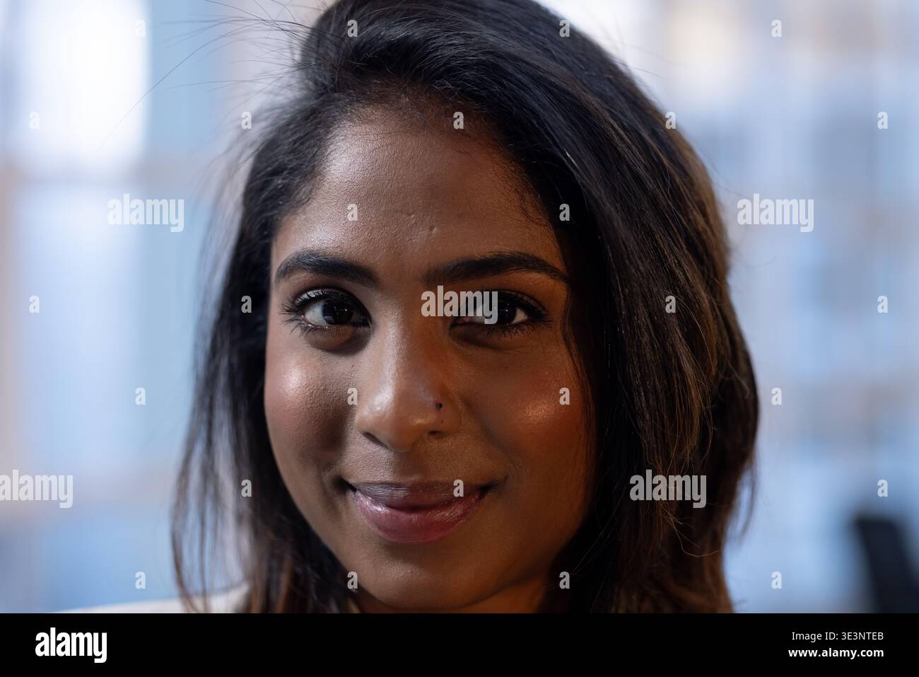 Donna indiana che sorride delicatamente in primo piano alla finestra dell'ufficio con il naso perforato e luce soffusa Foto Stock