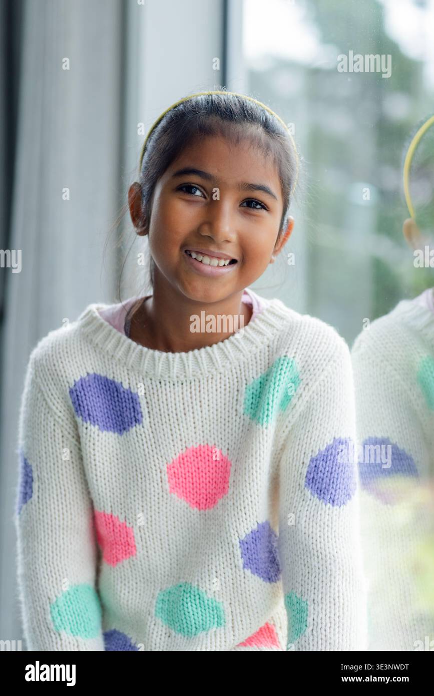 Ragazza asiatica sorridente, seduta a casa accanto alla finestra in maglione bianco con pois, fascia gialla Foto Stock