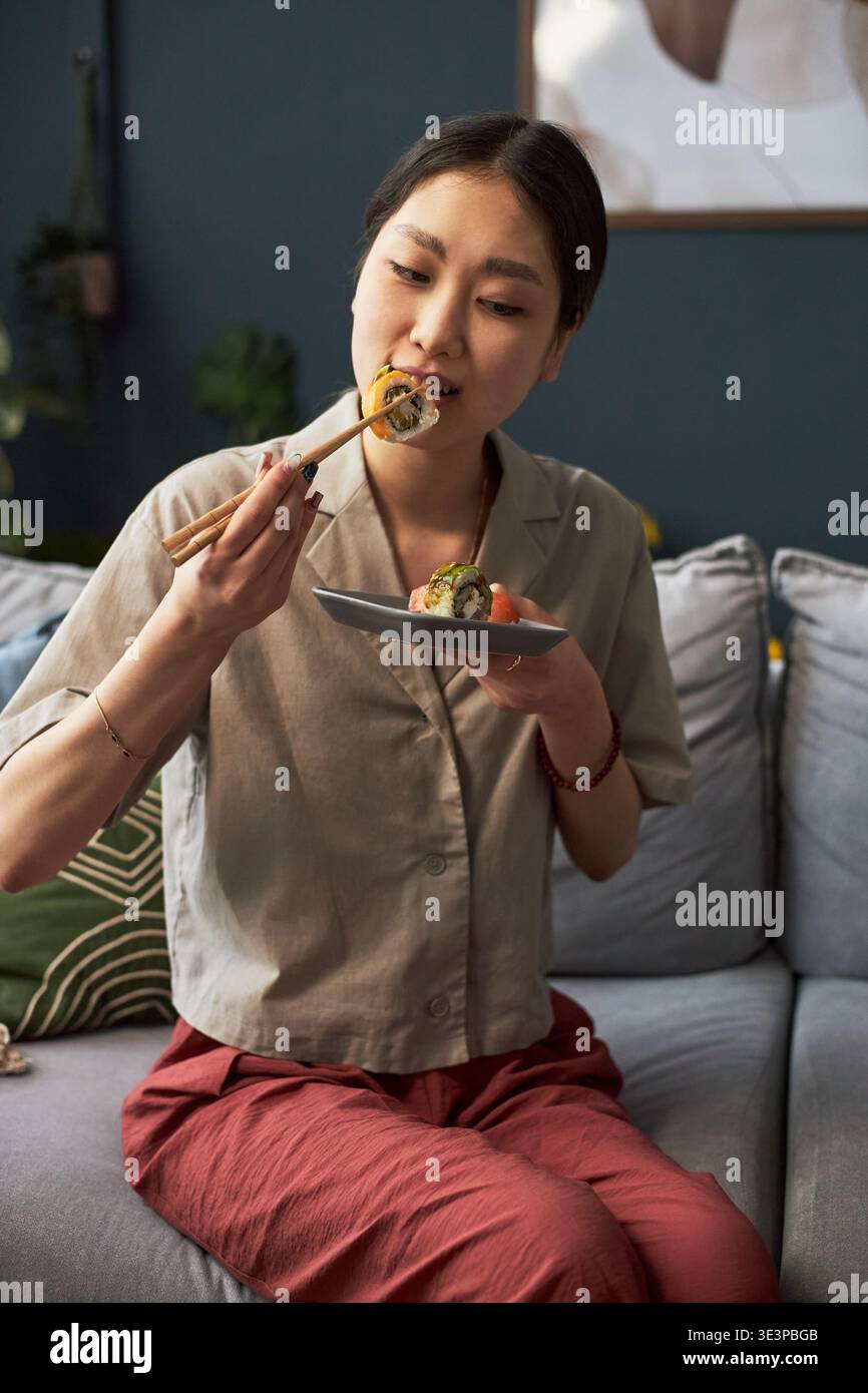 Giovane donna asiatica seduto sul divano che mangia sushi con bacchette che tengono in mano un piccolo piatto, mostrando la cultura culinaria tradizionale gustando un pasto in un ambiente familiare informale Foto Stock