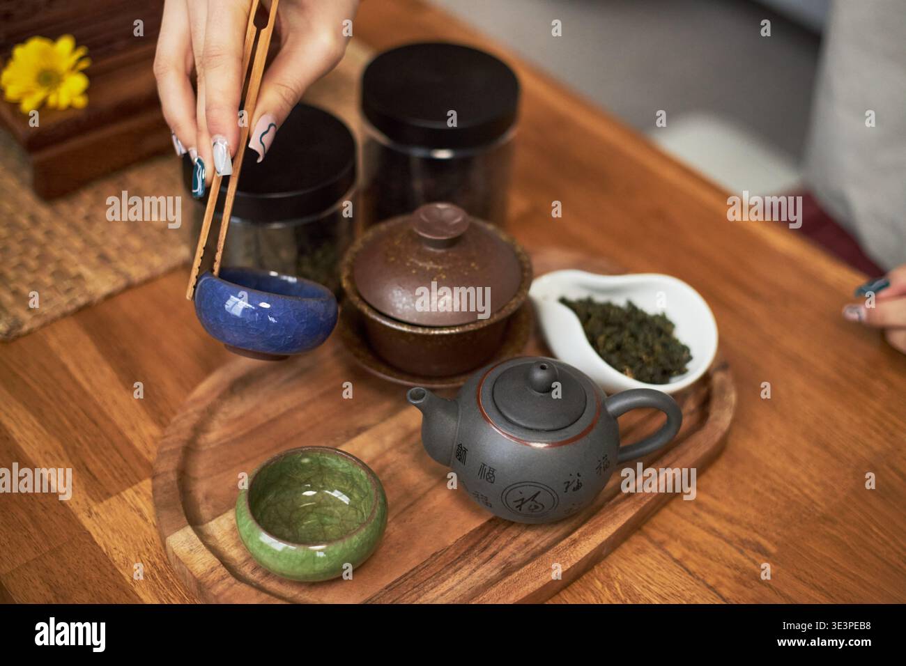 Giovane donna asiatica che prepara la tradizionale cerimonia del tè utilizzando bacchette di legno per gestire la tazza da tè in ceramica, le foglie di tè e la teiera disposte su un vassoio di legno che riflette il patrimonio culturale Foto Stock