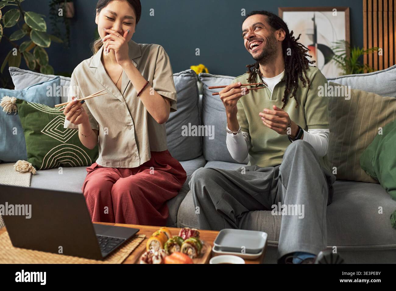 Giovane donna asiatica adulta e giovane uomo nero adulto seduto sul divano ridendo e mangiando con le bacchette mentre guardano il portatile, condividendo momenti di gioia e gustando insieme la cucina asiatica Foto Stock