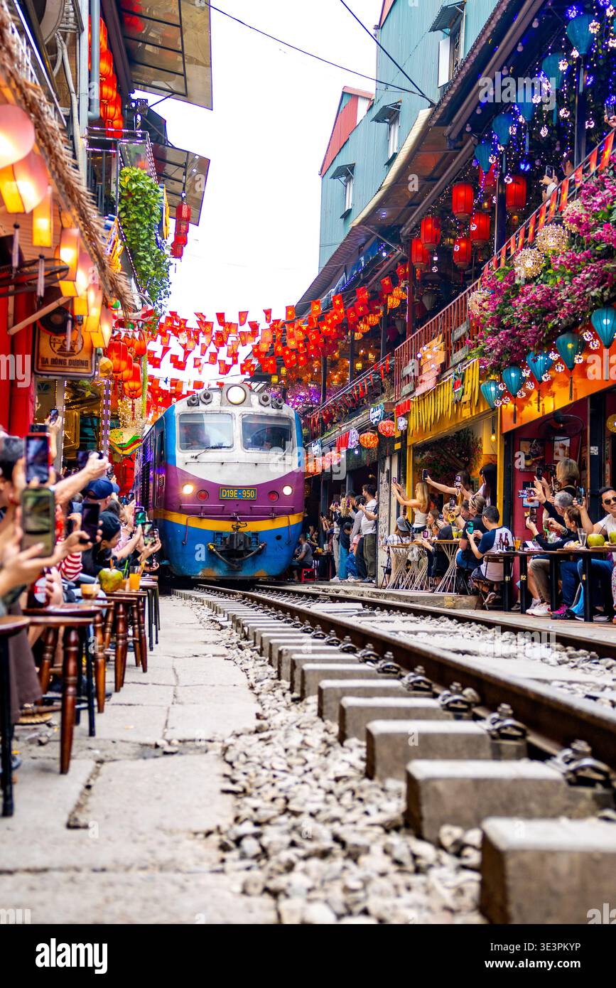 Hanoi, Vietnam - 19 gennaio 2026: Vista della vivace via dei treni, dove un treno blu e viola attraversa la vivace corsia adornata di colori Foto Stock
