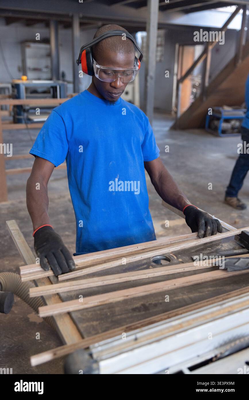 Uomo afroamericano che maneggia strisce di legno sul tavolo dell'officina indossando occhiali di sicurezza e cuffie Foto Stock