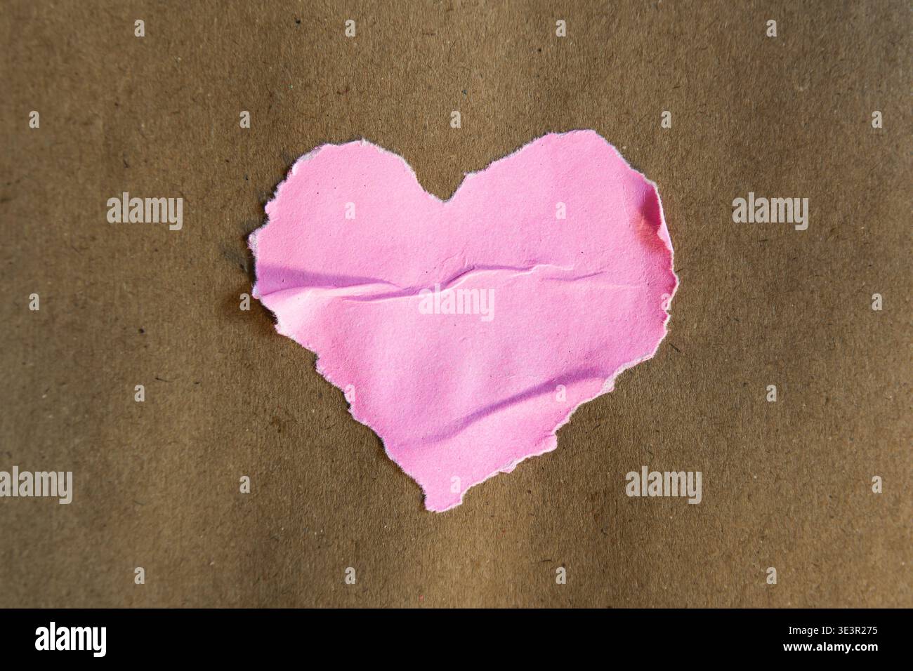 Forma a cuore rosa fatta a mano con bordi strappati e irregolari incollati al centro di uno sfondo rustico di carta marrone. Lo shot cattura il bordo fibroso tattile Foto Stock