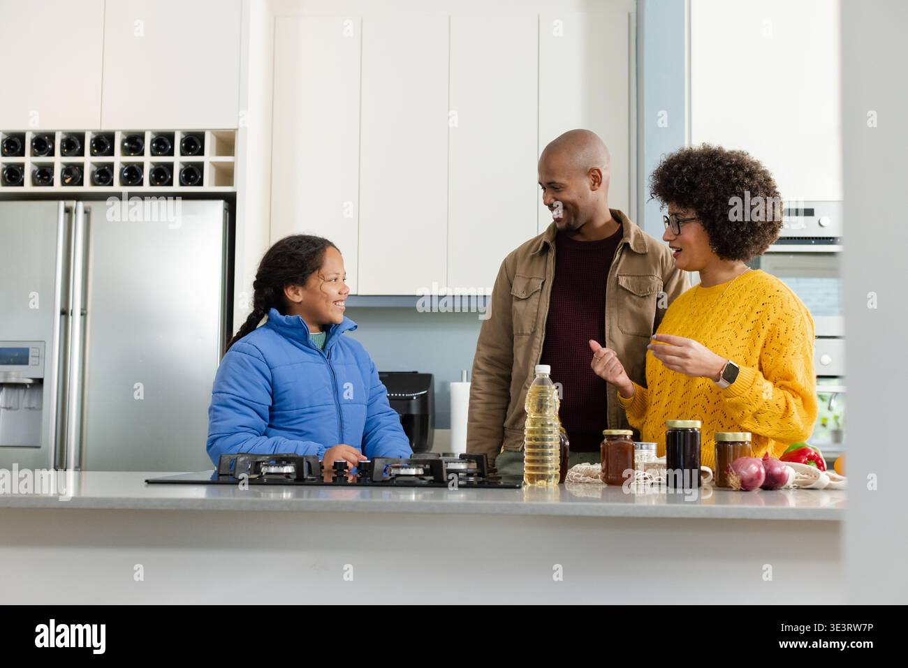 Famiglia diversificata che si trova a Kitchen Island per preparare i pasti utilizzando una stufa a gas integrata e vasetti di spezie Foto Stock