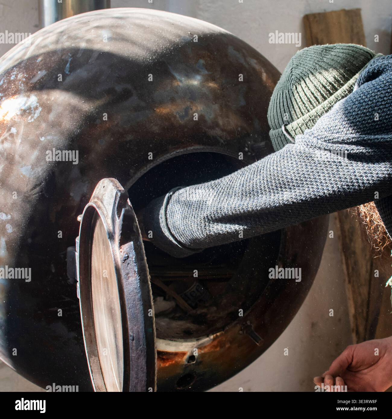 Artigiano barbuti che accende una stufa in metallo fatta a mano all'interno di un laboratorio di lavorazione del legno. Area di lavoro industriale con apparecchiature di riscaldamento utilizzate in un ambiente rustico Foto Stock