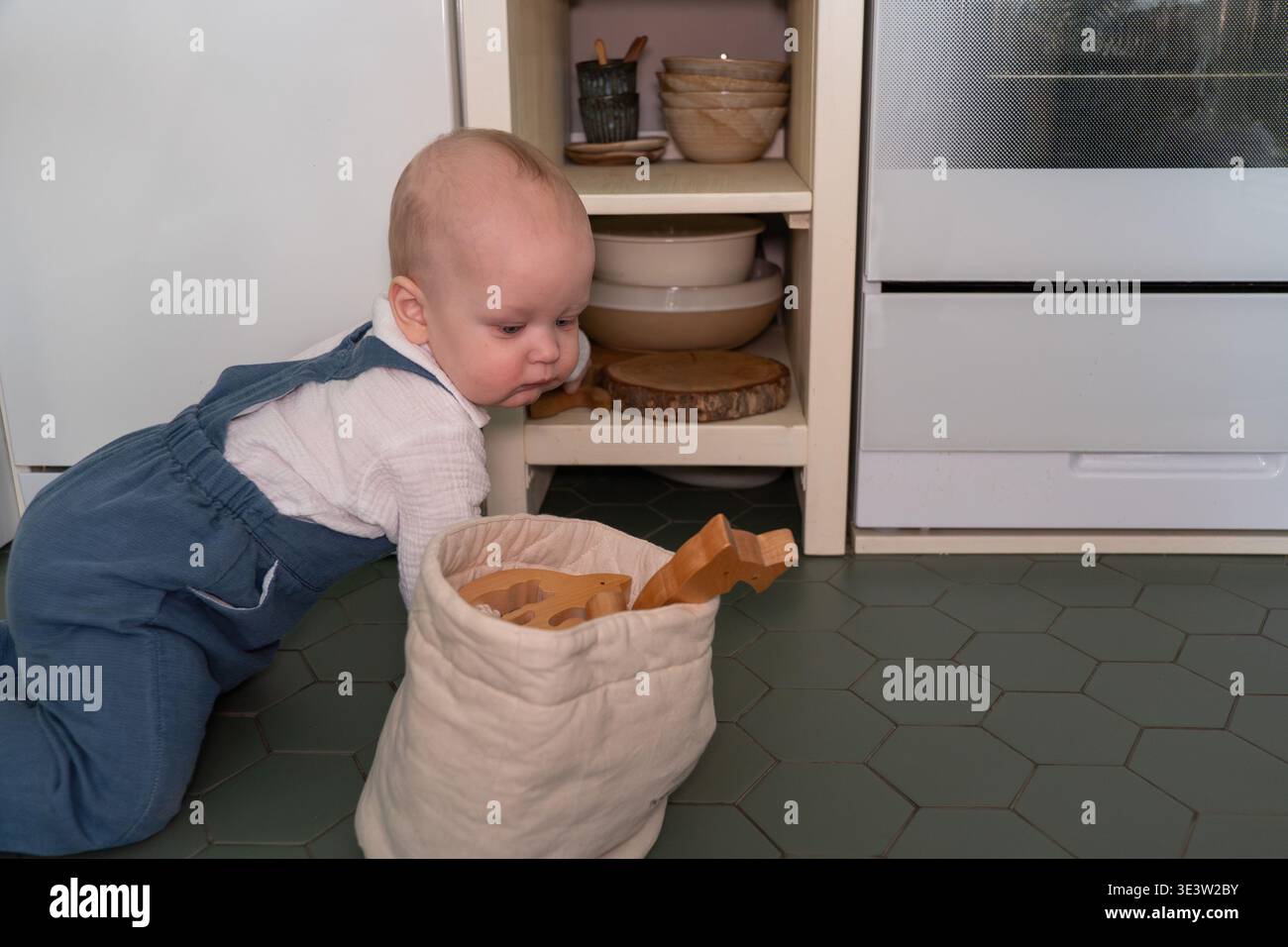 Il bambino si inginocchia sul pavimento della cucina entrando in una borsa in tessuto con giocattoli in legno negli interni della casa, esplorazioni quotidiane e momenti di gioco. Sviluppo del bambino Foto Stock