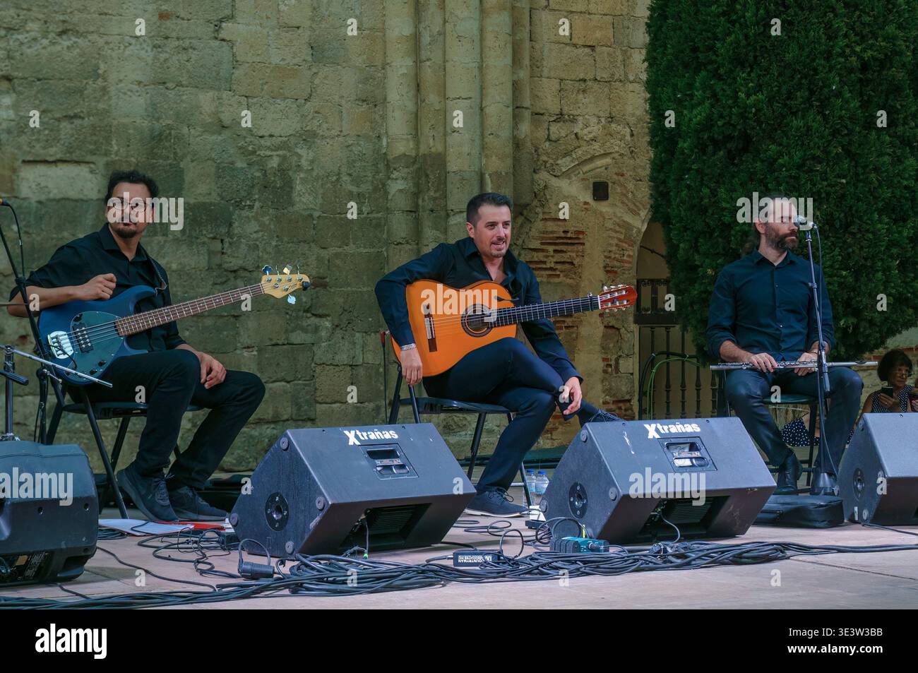 Tre musicisti maschi che si esibiscono dal vivo su un palco a Valladolid, Spagna, con chitarra, basso e flauto su uno sfondo storico in pietra. Foto Stock