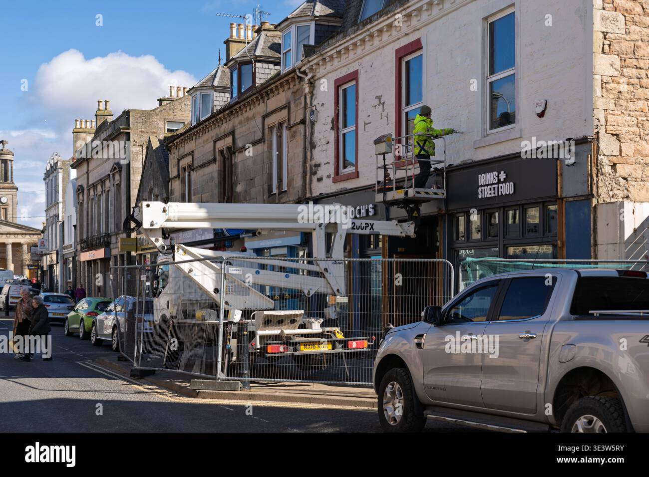 Un lavoratore con attrezzatura ad alta visibilità utilizza una piattaforma sopraelevata per raschiare la parete esterna di un edificio di Drinks Street Food a Elgin, Moray, Scozia. Le auto parcheggiate e le persone si spostano lungo la strada soleggiata. Crediti: JASPERIMAGE/Alamy Live News Foto Stock