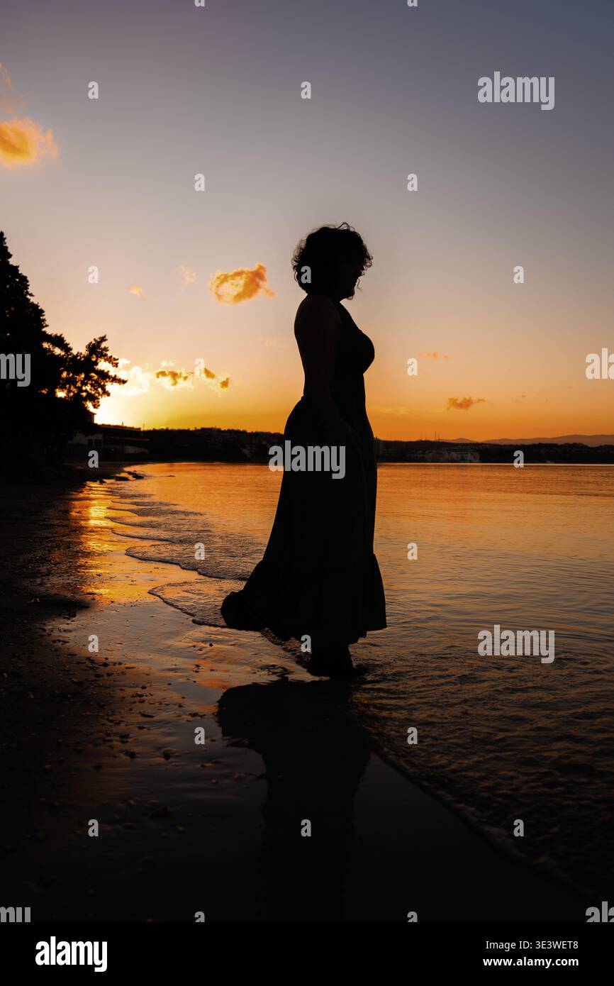 Silhouette di una donna in un abito fluente che cammina sulla spiaggia durante un vibrante tramonto. Il cielo è dipinto in sfumature d'arancio e d'oro, riflettendo Foto Stock