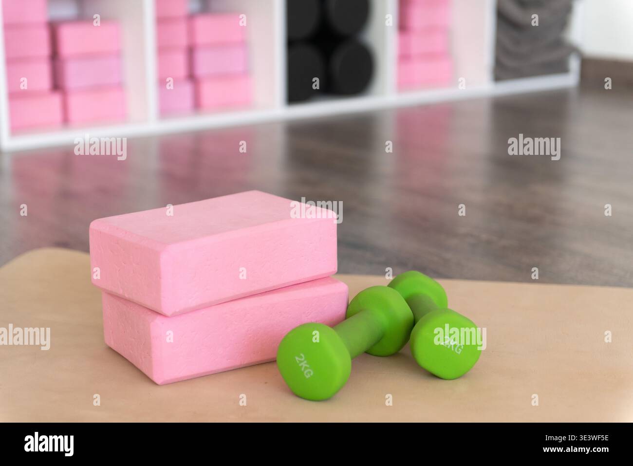 Un set di blocchi di yoga rosa e due manubri verdi da 2 kg sono disposti su un pavimento di legno della palestra, con scaffali che contengono attrezzature fitness aggiuntive nella ba Foto Stock
