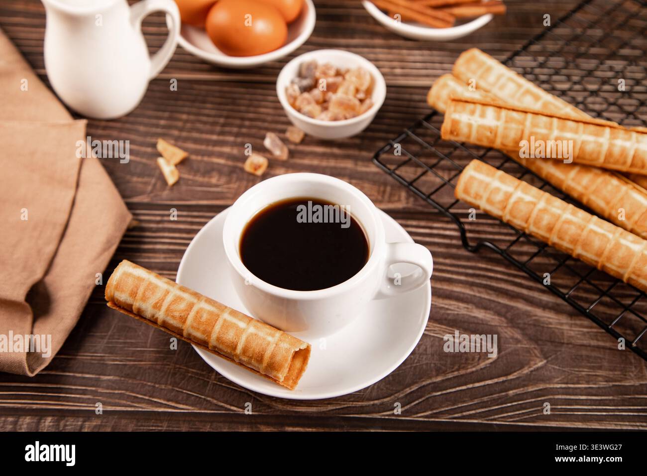 Una tazza di caffè nero fumante è abbinata a biscotti croccanti arrotolati su un tavolo in legno rustico. Ingredienti come uova, zucchero e cannella aggiungono una h Foto Stock