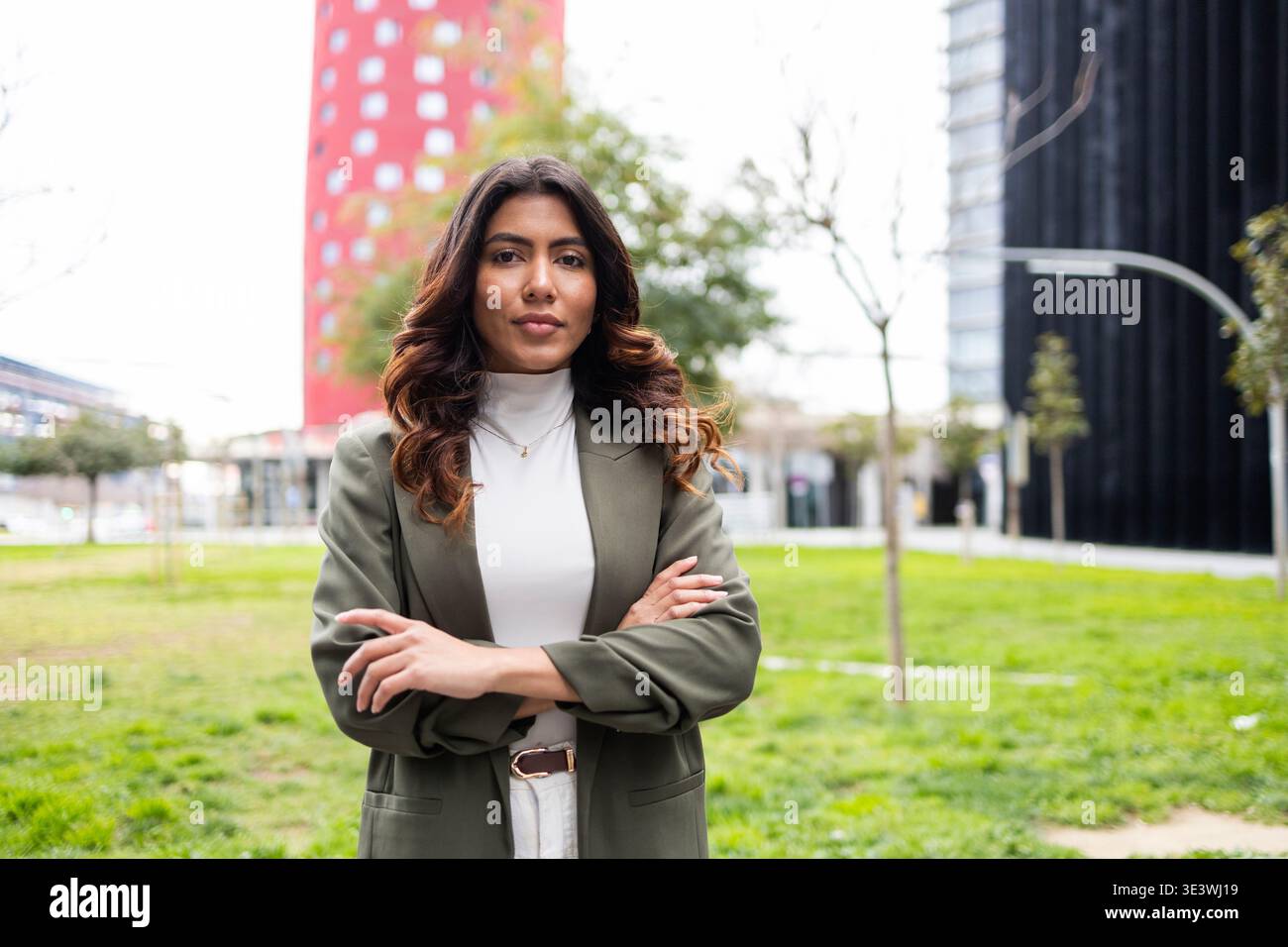 Donna d'affari professionista in un blazer verde si trova in un moderno parco urbano. Lo sfondo presenta edifici contemporanei e lussureggiante greene Foto Stock