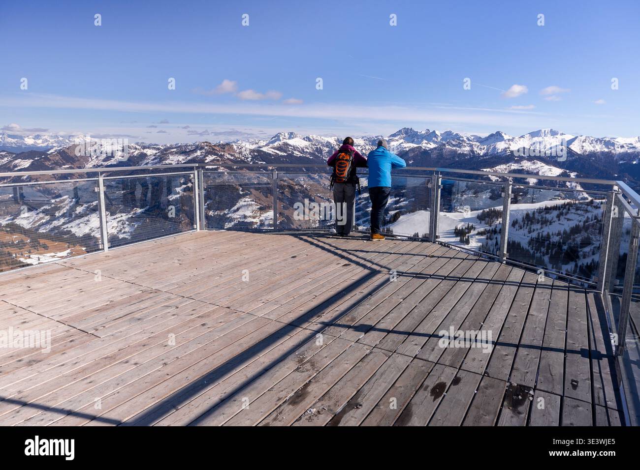 Due persone godono di una vista invernale mozzafiato da una piattaforma di osservazione in legno sulle Alpi. Le montagne innevate si estendono maestosamente sotto un blu chiaro Foto Stock