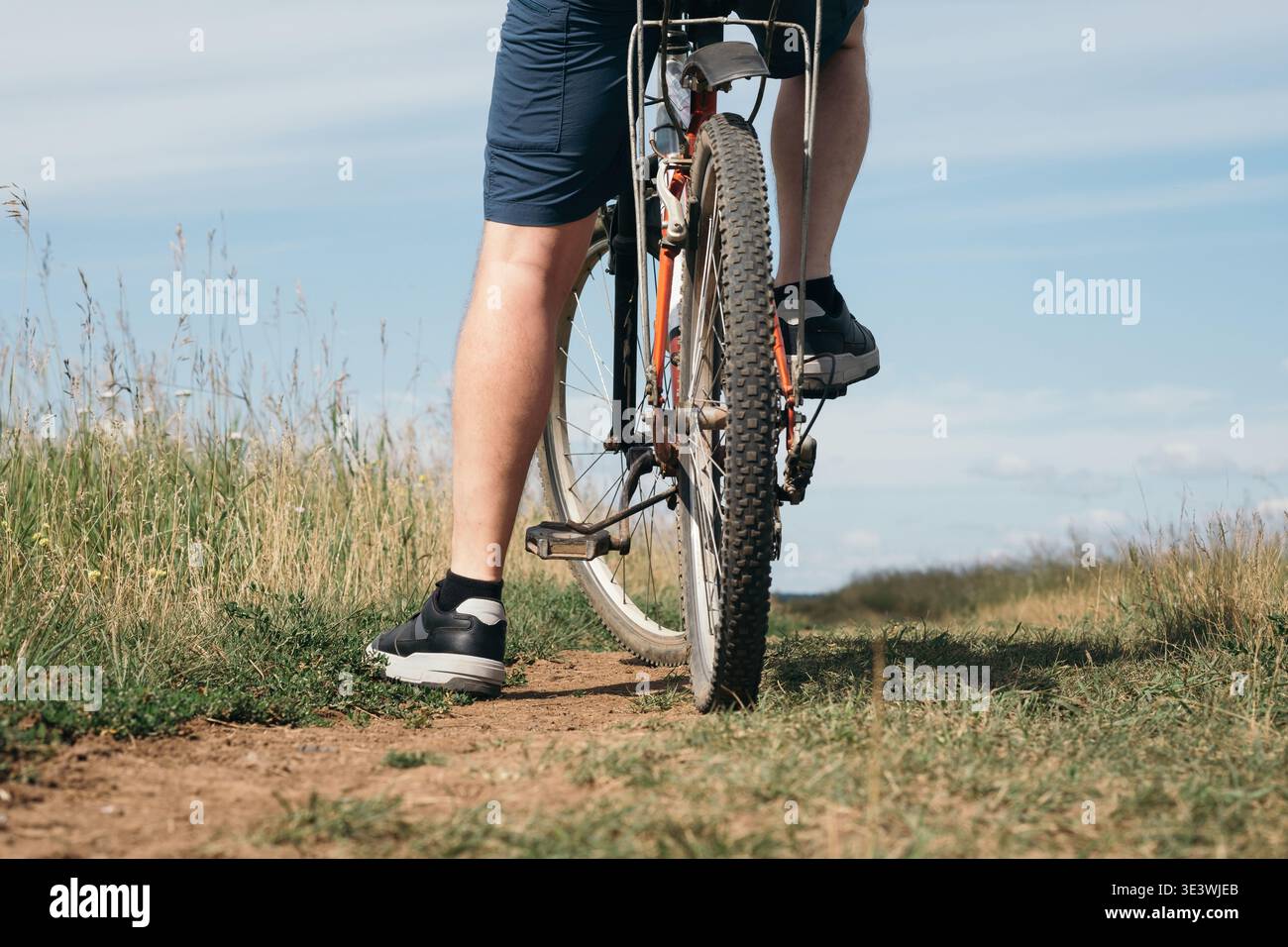 Un ciclista che indossa pantaloncini corta percorre un'aspra mountain bike lungo un sentiero sterrato. Il percorso è fiancheggiato da erba alta sotto un cielo blu limpido, catturando un momento Foto Stock