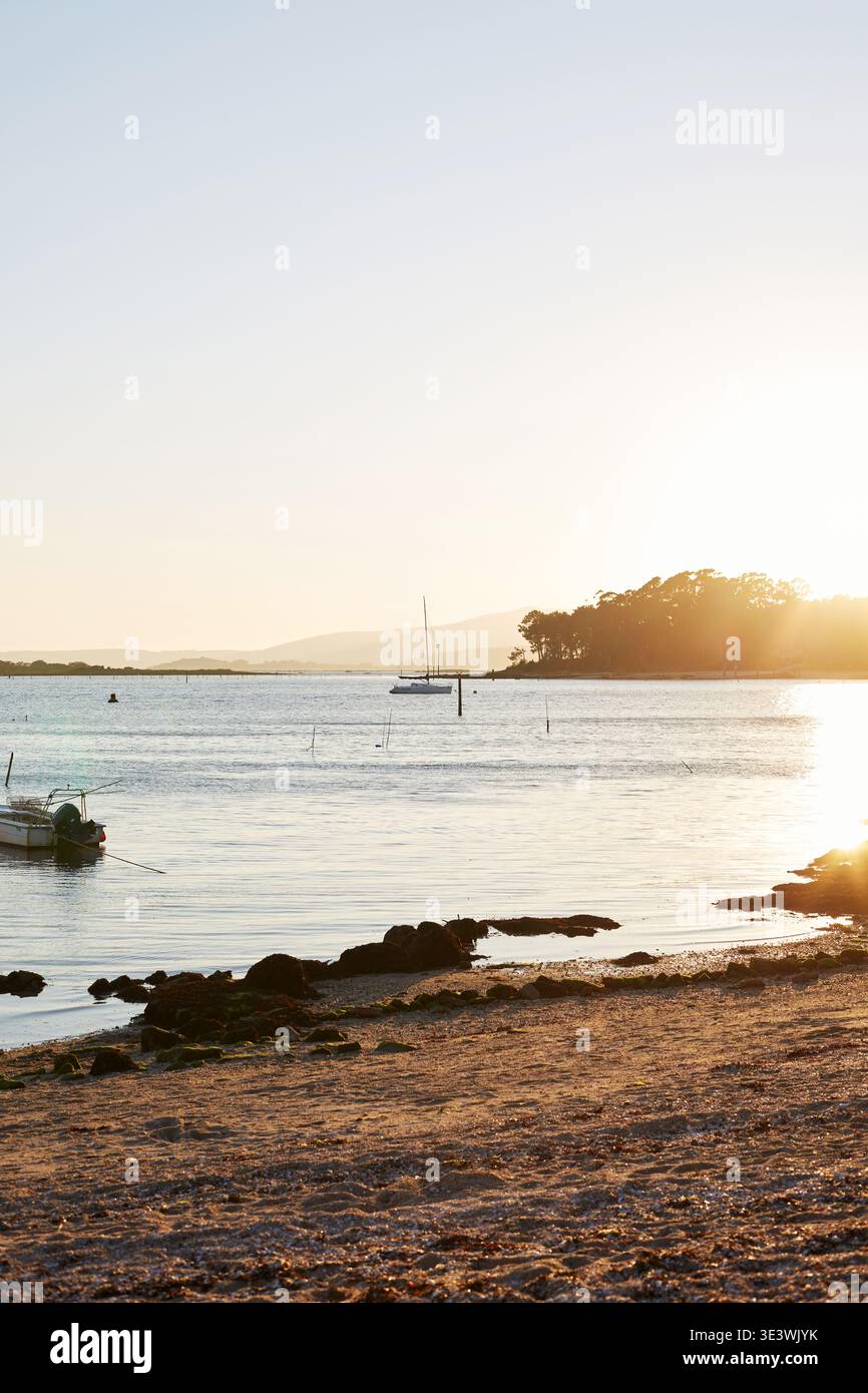 Serena vista serale della Galizia, Spagna, con il sole che tramonta sulle acque calme e le coste rocciose. Uno yacht galleggia in lontananza, creando un ambiente tranquillo Foto Stock