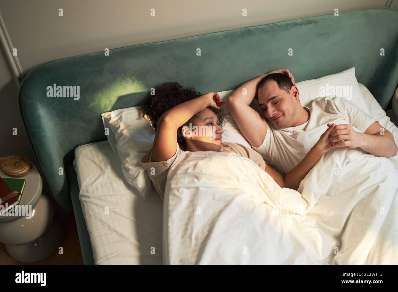 Giovane donna nera adulta e giovane uomo caucasico adulto sdraiati a letto insieme sotto la coperta, di fronte l'un l'altro e sorridente, rilassato momento mattutino in camera da letto Foto Stock