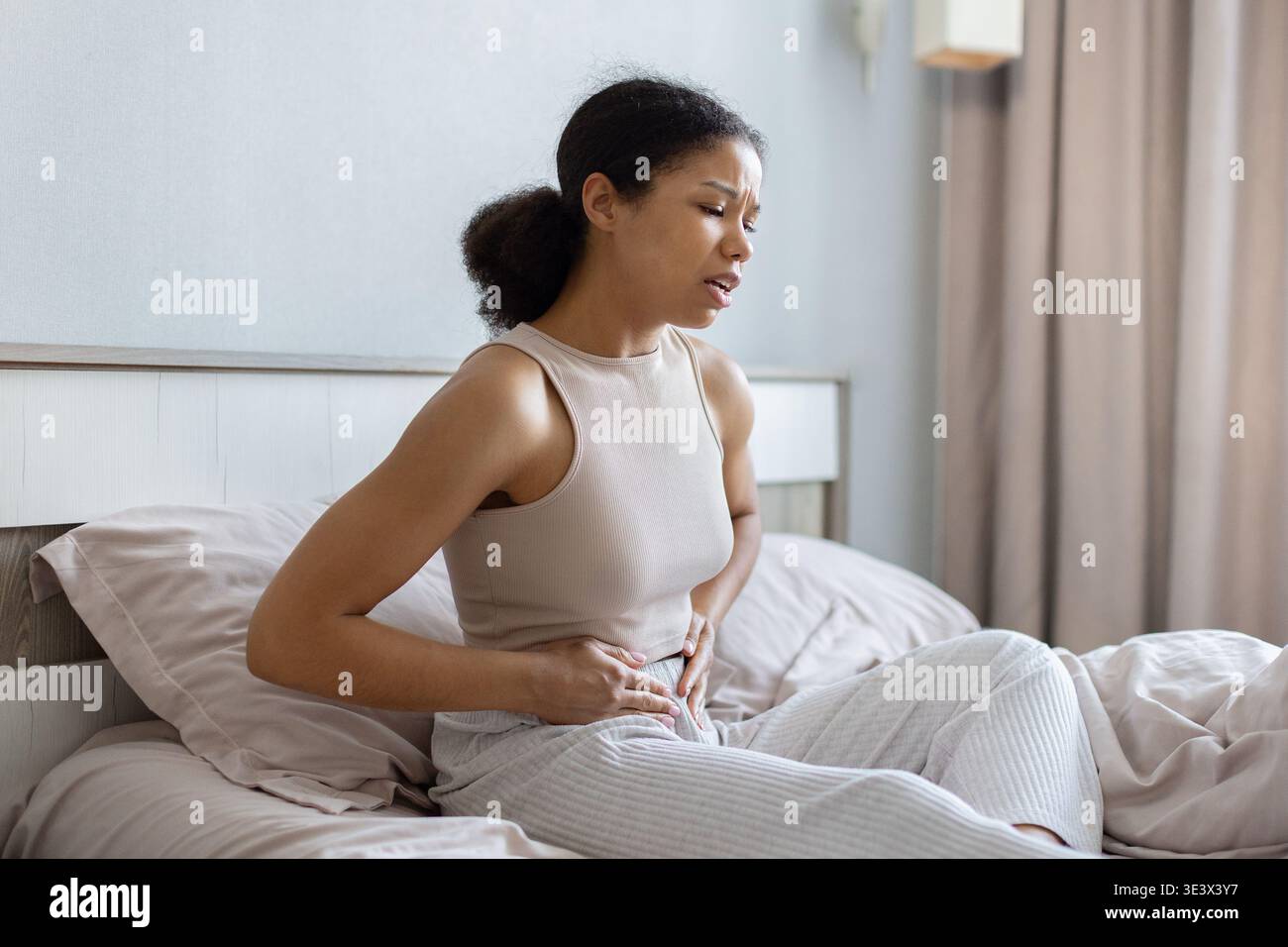 Donna nera seduta sul letto che tiene l'addome basso nel dolore, mostrando disagio Foto Stock