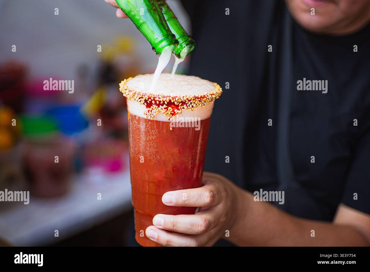 Persona che versa due birre contemporaneamente in un bicchiere michelada preparato con peperoncino, semi di sesamo e bordo di salsa camoscio. Foto Stock
