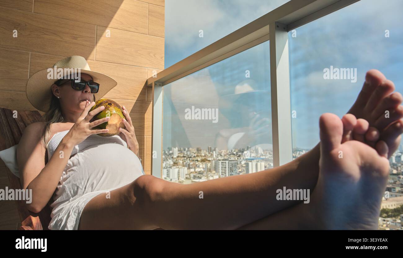 Donna incinta che si gode un drink tropicale e vista della città da un balcone soleggiato Foto Stock