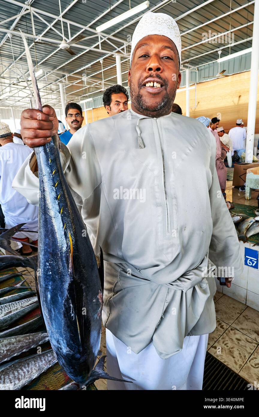 Un uomo si trova nel mercato del pesce di Mascate, Oman, con un pesce grande. Altre persone navigano nelle vicinanze. Il mercato è affollato e vivace. Foto Stock
