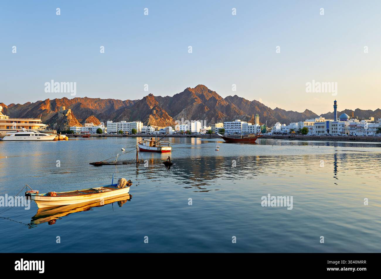 Le onde riflettono la luce del tramonto a Mascate, Oman, mentre le barche si trovano in acqua sulla riva. Le montagne sorgono dietro edifici in lontananza. Foto Stock