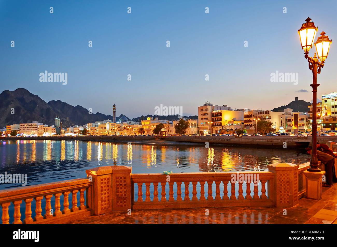 Il tramonto proietta un bagliore sul lungomare di Mascate, Oman. Le luci degli edifici si riflettono nelle acque calme, creando un'atmosfera vivace. Foto Stock