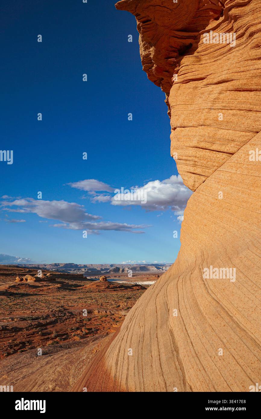 Formazione di roccia arenaria curva che si affaccia sul terreno desertico con mesa lontane e cielo blu con nuvole sparse in un paesaggio arido Foto Stock
