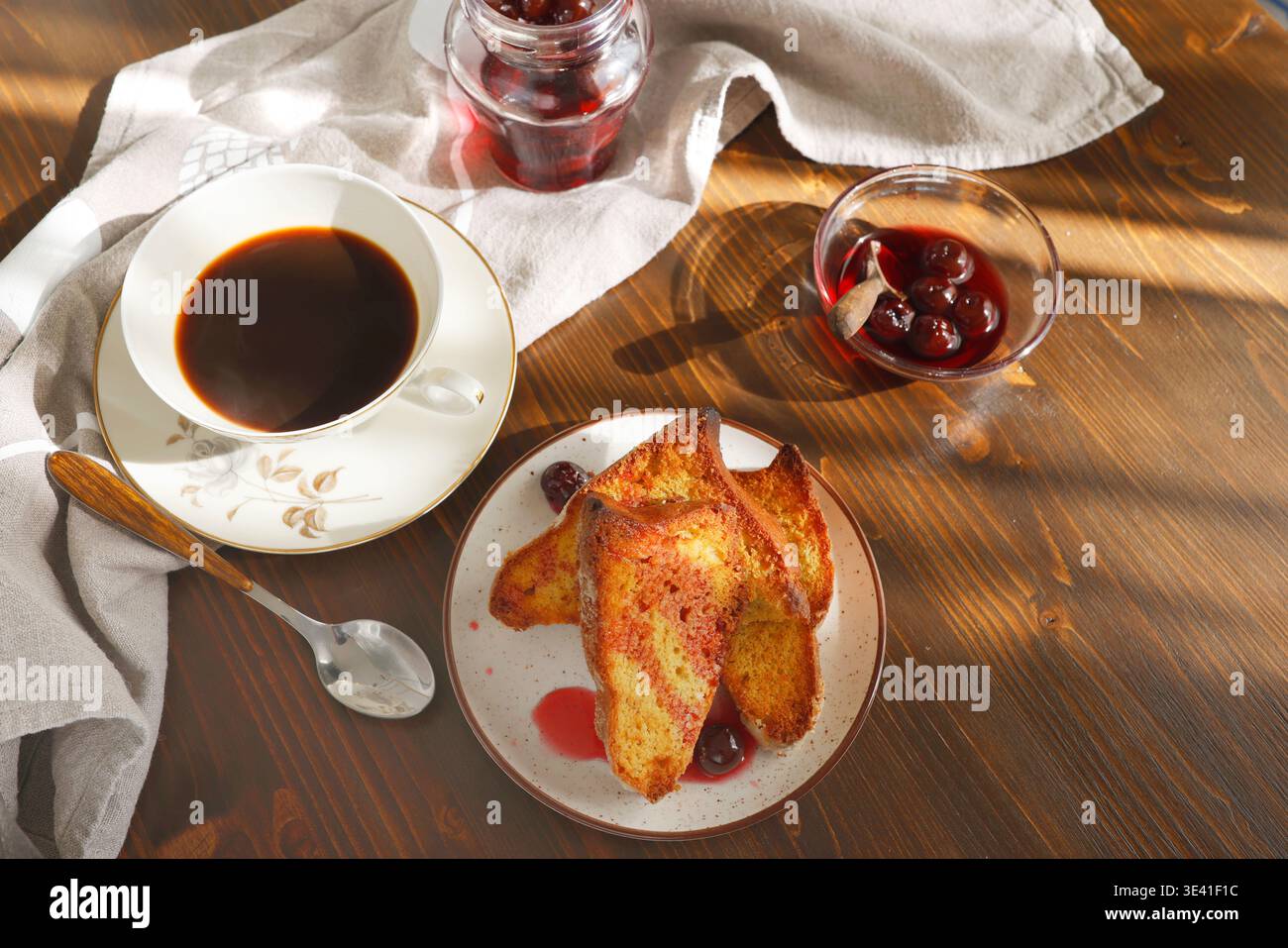 Toast alla francese con marmellata di ciliegie e tazza di caffè nero su un tavolo di legno. Le fette di brioche dorate vengono servite con sciroppo di bacca e espresso per una colazione accogliente Foto Stock