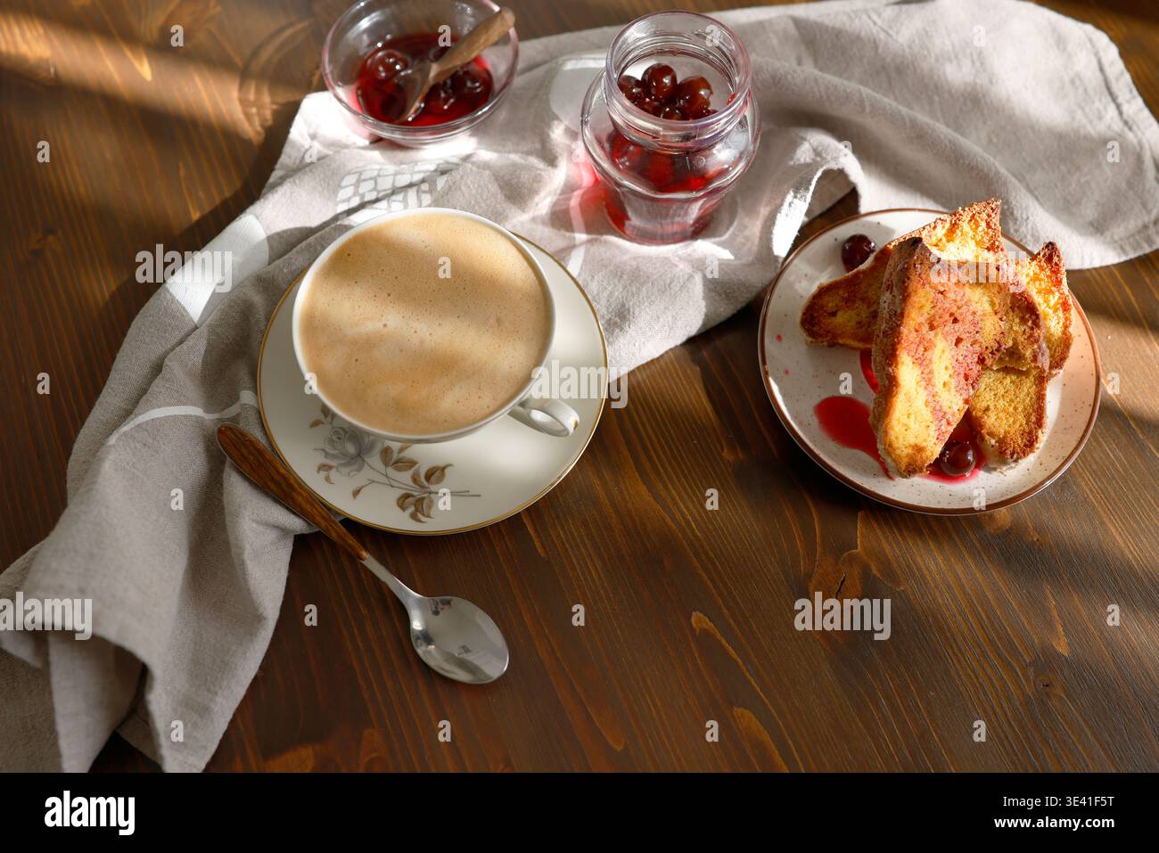Toast alla francese con marmellata di ciliegie e tazza di cappuccino su un tavolo di legno. Le fette di brioche dorate sono servite con sciroppo di bacca e caffè schiumoso per una colazione accogliente Foto Stock