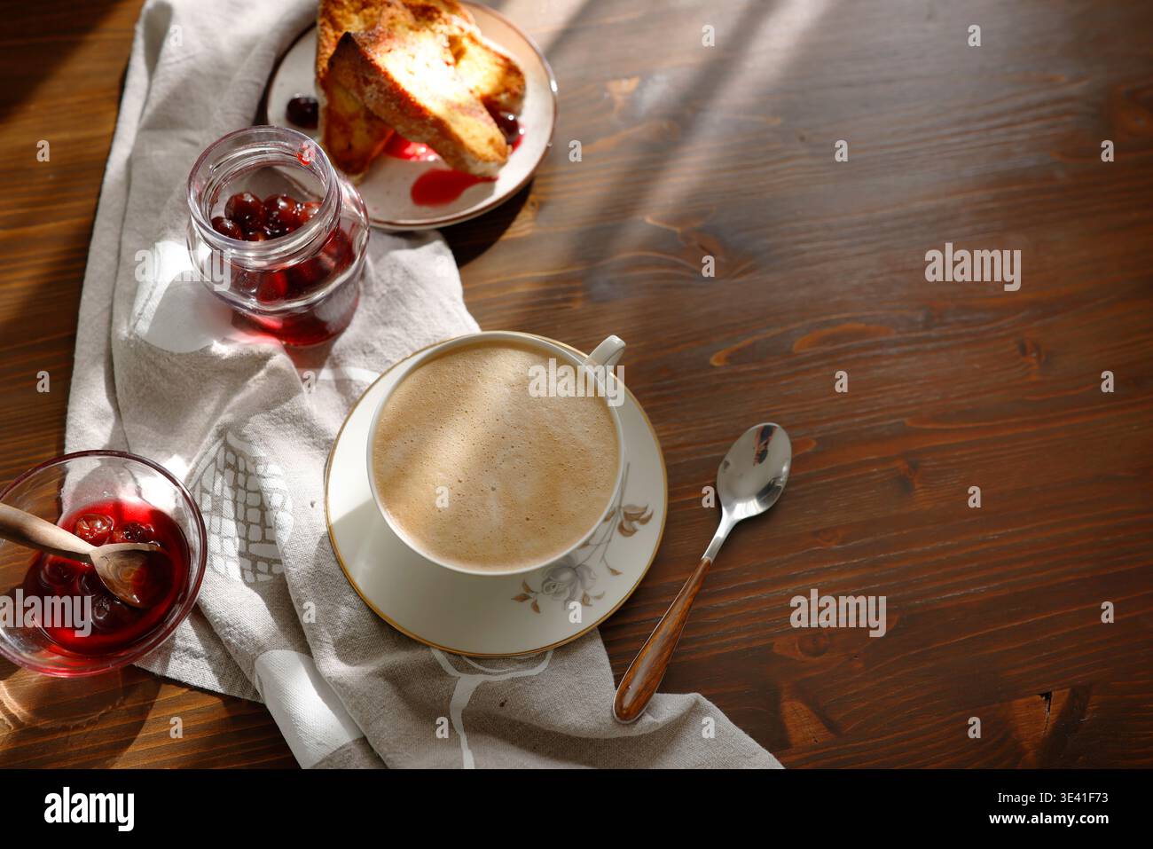 Toast alla francese con marmellata di ciliegie e tazza di cappuccino su un tavolo di legno. Le fette di brioche dorate sono servite con sciroppo di bacca e caffè schiumoso per una colazione accogliente Foto Stock