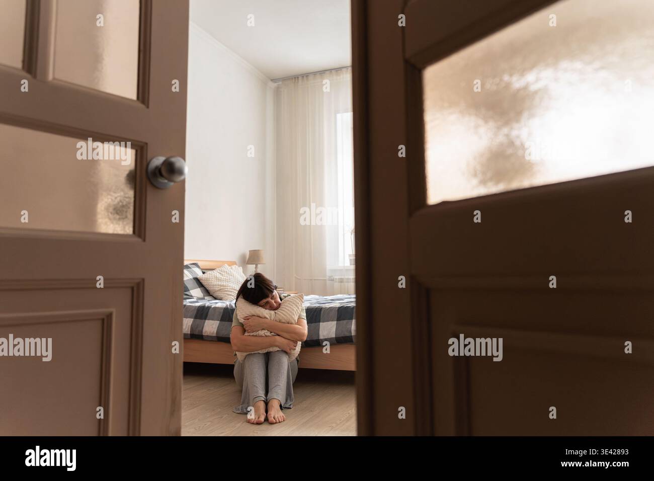 Donna che abbraccia il cuscino mentre siede sul pavimento della camera da letto, si sente sola, triste e soffre di problemi di salute mentale Foto Stock