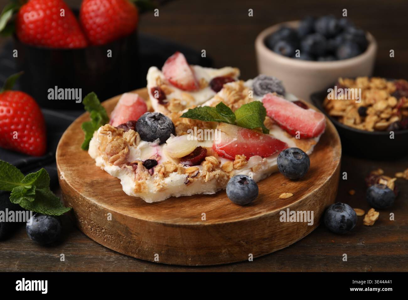 Pezzi di gustosa corteccia di yogurt con frutti di bosco, menta e muesli su tavola di legno, primo piano Foto Stock