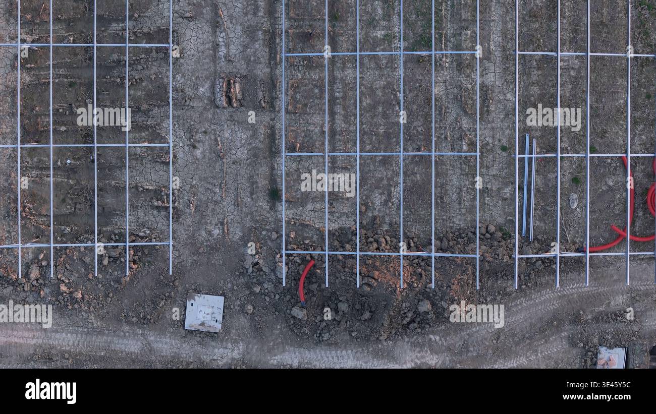 Predisposizione per la messa a terra per l'installazione di pannelli fotovoltaici, con telai dei moduli in metallo e condotti elettrici rossi su terra sterile, a indicare il progresso Foto Stock