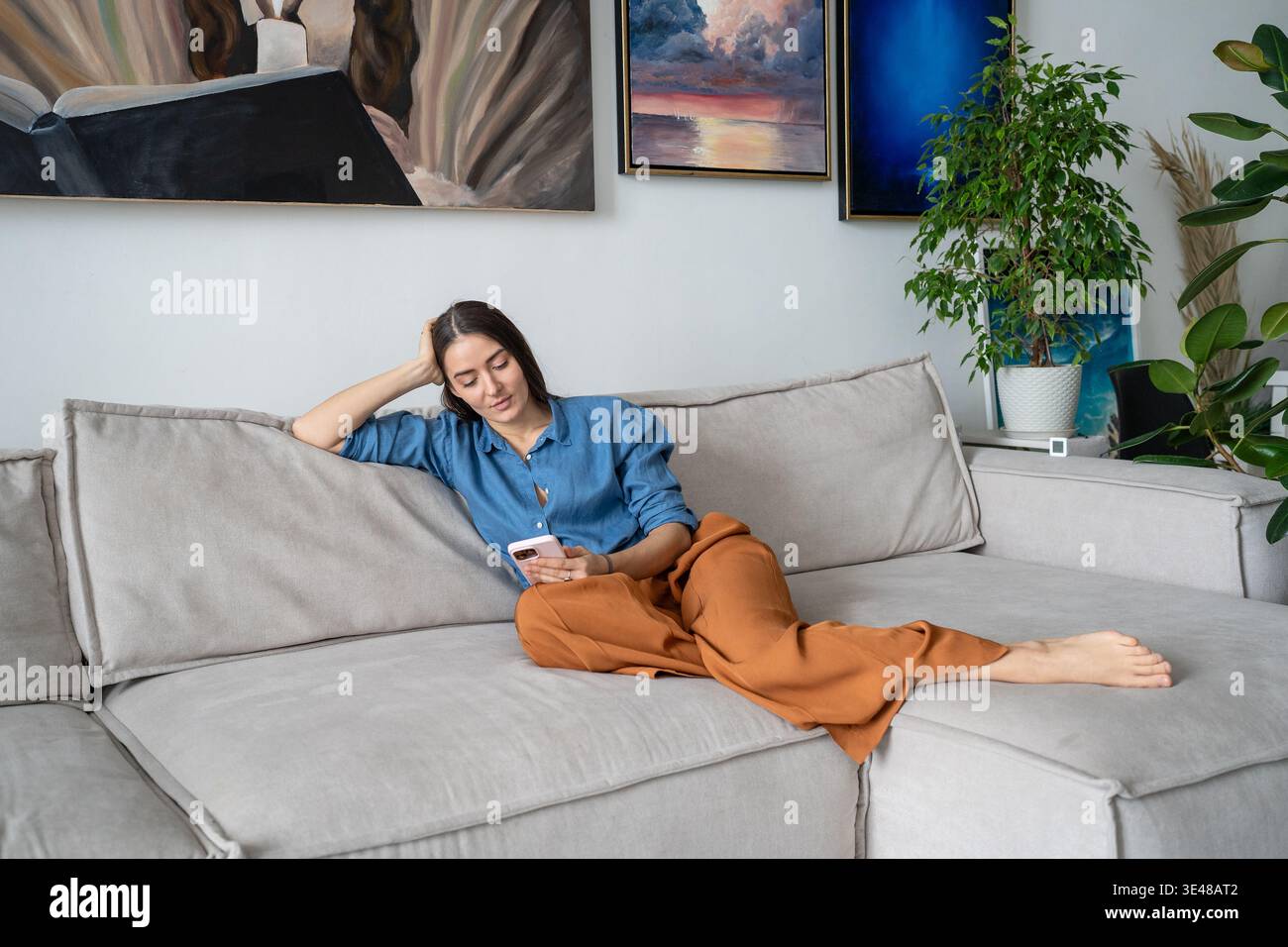 Donna sorridente e calma, pittrice che si rilassa sul divano procrastinando i contenuti multimediali a scorrimento temporale sullo smartphone. Foto Stock