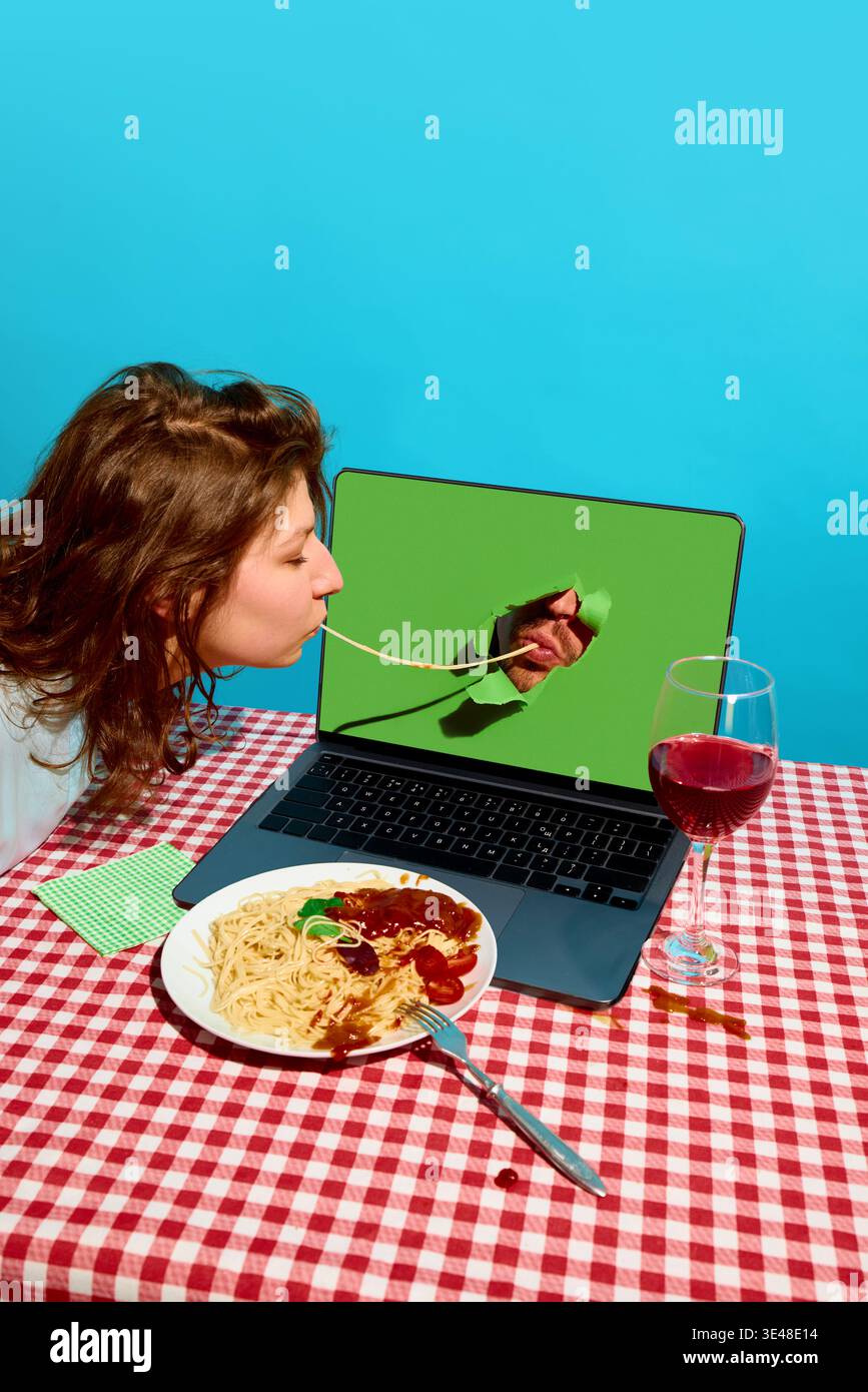 Donna che mangia spaghetti collegati all'uomo attraverso lo schermo del laptop a tavola con vino. Foto Stock