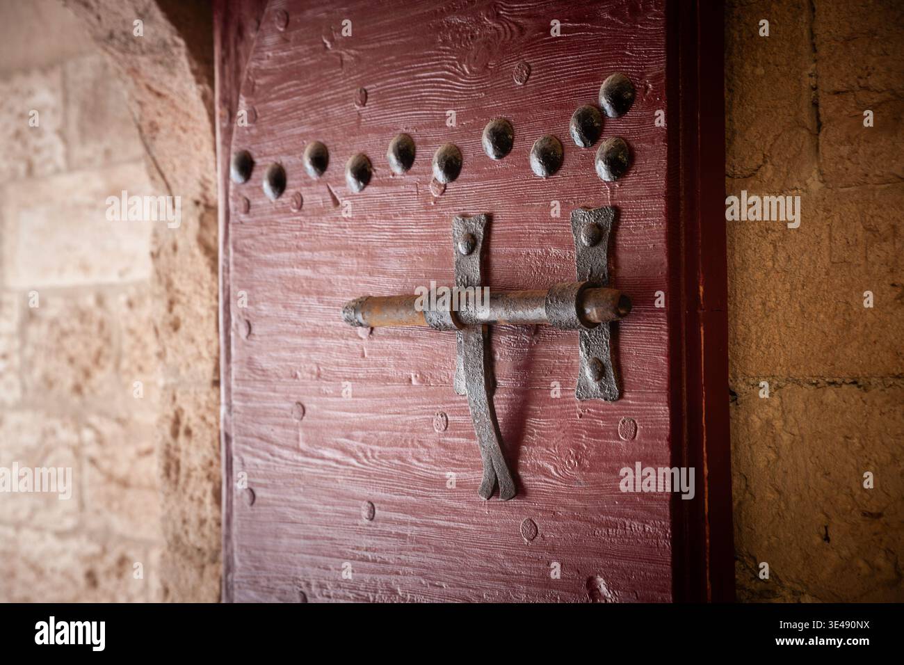Primo piano di una serratura di ferro su una porta in legno rossa con borchie decorative in metallo e pietre circostanti, sottolineando la consistenza rustica e i vecchi oggetti di artigianato Foto Stock