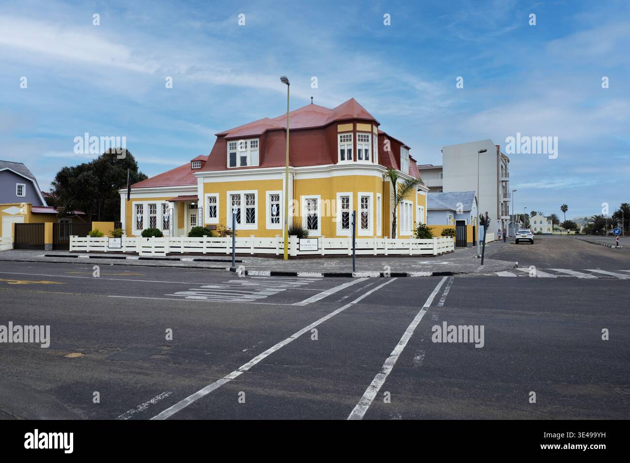 Vista di un edificio giallo luminoso in stile coloniale con un tetto rosso su un angolo di strada con una recinzione bianca sotto un cielo blu a Swakopmund, regione di Erongo, N Foto Stock