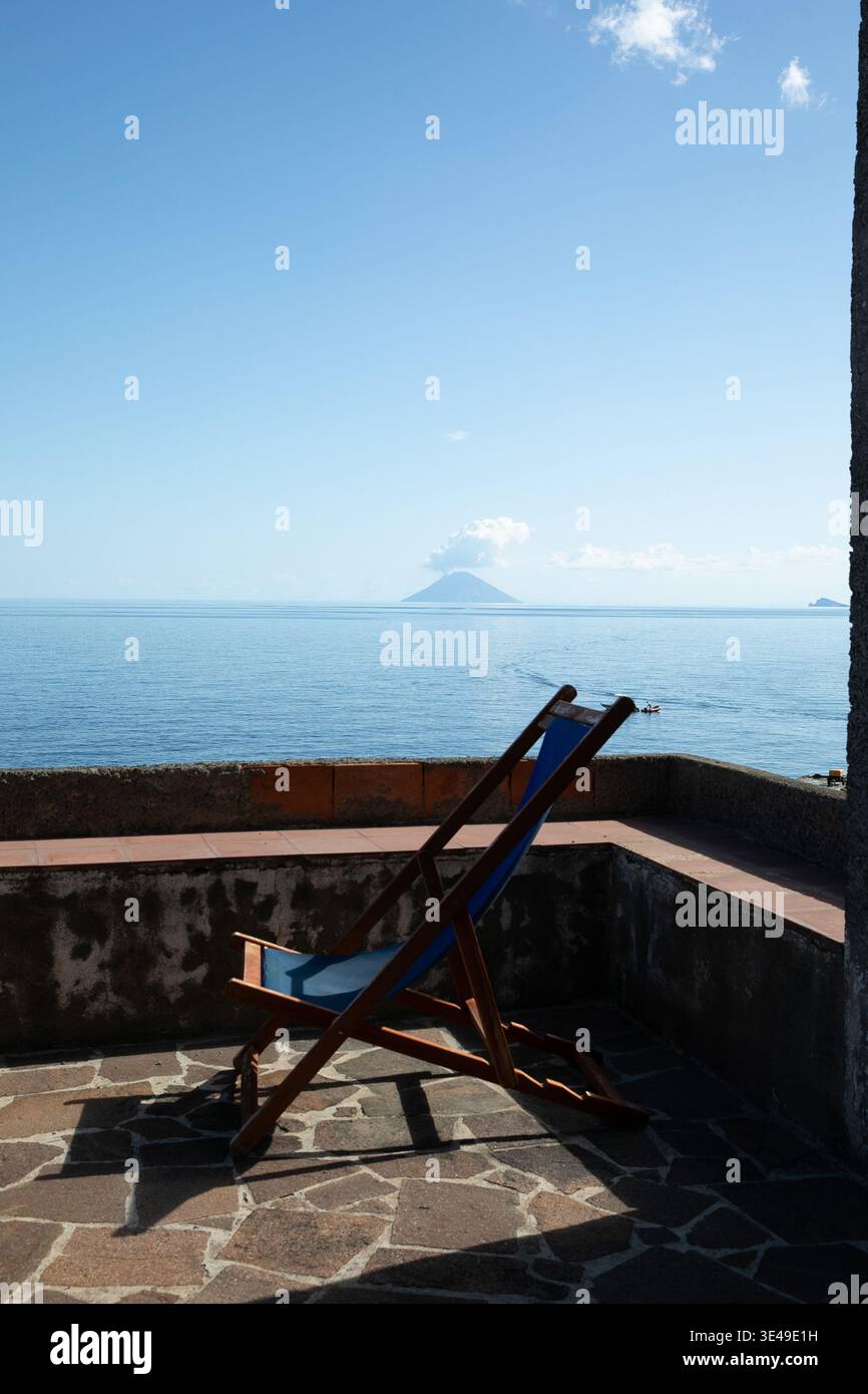 Vista di una sedia a sdraio blu su una terrazza in pietra che si affaccia sul Mar Tirreno con il vulcano Stromboli fumante all'orizzonte sotto un cielo limpido a Sal Foto Stock
